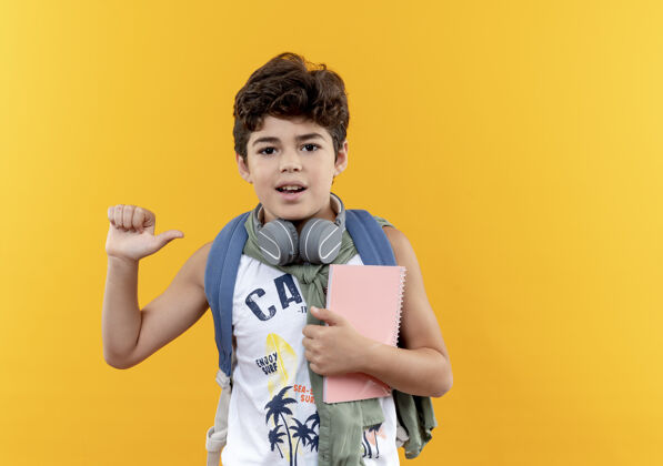 请戴上高兴的小男生背着书包戴着耳机拿着笔记本隔离在黄色背景上拿着耳机小的