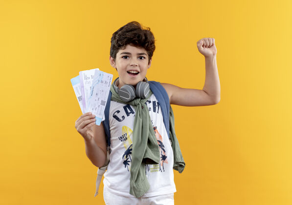 穿快乐的小男孩戴着背包和耳机拿着票 在黄色背景上显示“是”的手势抱小包