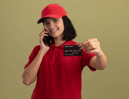 年轻人身穿红色制服 戴着帽子的年轻送货员在讲手机 信用卡微笑着站在轻墙上欢呼站立表演