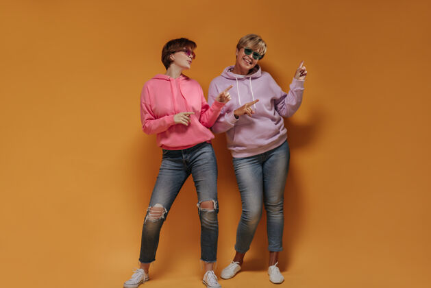 中等两位短发女士的全幅照片 戴着明亮的太阳镜 淡紫色和粉色的连帽衫 牛仔裤 在橙色的背景上展示文字的位置女老年人衬衫