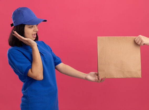 年轻穿着蓝色制服 戴着帽子的年轻送货女孩站在粉色的墙上 一边接受纸包裹 一边兴奋不已制服女孩兴奋