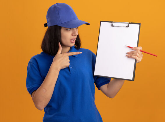 显示身穿蓝色制服 戴着帽子的年轻送货员站在橙色的墙上 手里拿着空白页和铅笔 用食指指着他们剪贴板制服手指