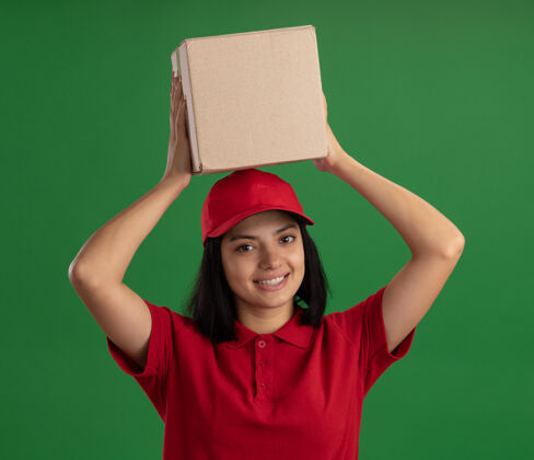 立场身穿红色制服 头戴鸭舌帽 头上抱着纸板箱 友好微笑地站在绿色墙壁上的年轻送货女孩年轻微笑制服