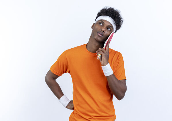 乒乓球看着一边思考的年轻的美国黑人运动男子戴着头带和腕带把乒乓球拍放在脸颊上孤立的白色背景脸颊运动球拍