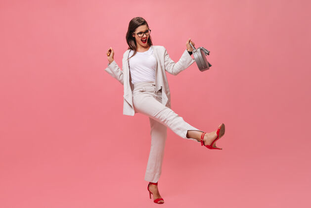 夹克穿西装的活跃女士在粉色背景上拿着手提包摆姿势情绪化的年轻女子穿着红色高跟鞋在镜头前玩得很开心高跟鞋包白色
