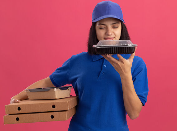 年轻人身穿蓝色制服 头戴鸭舌帽 拿着比萨饼盒和食品包的年轻送货女孩站在粉色的墙上 看上去自信而快乐制服帽子盒子