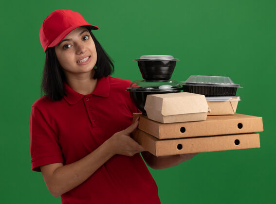 盒子身穿红色制服 头戴鸭舌帽 手持比萨饼盒和食品包的年轻送货女孩站在绿色的墙上 脸上带着困惑的微笑食物送货年轻