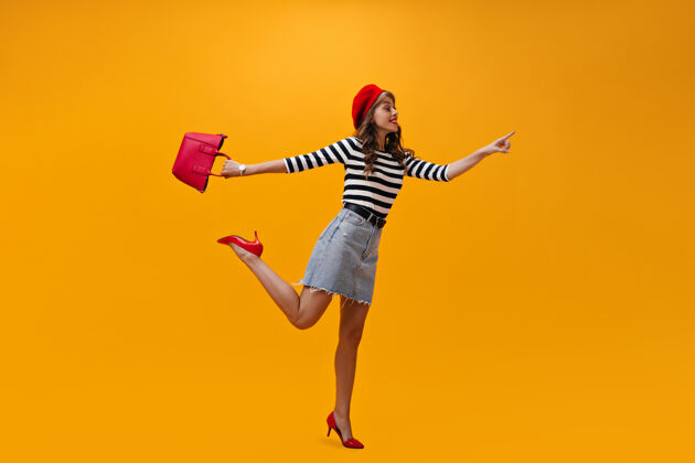 微笑兴高采烈的女人指着橙色背景上的文字微笑着穿着条纹毛衣和红色高跟鞋的时髦女孩摆姿势成人女孩帽子