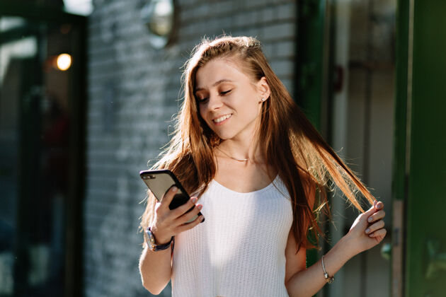 现代快乐美丽的年轻女子 留着浅棕色的长发 在阳光明媚的街道上用智能手机玩头发成人模特午餐