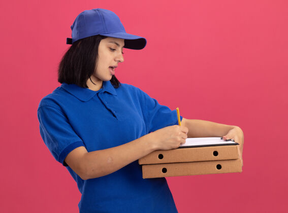 披萨穿着蓝色制服 戴着帽子的年轻送货女孩拿着比萨饼盒和剪贴板 站在粉红色的墙上严肃地写着什么拿着剪贴板盒子