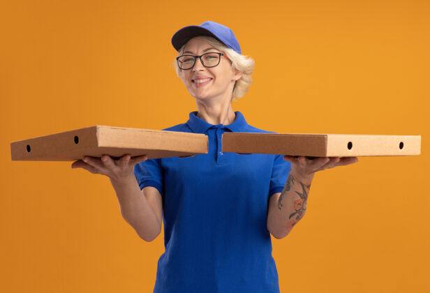 盒子身着蓝色制服 戴着眼镜 脸上挂着大大笑容 手拿比萨饼盒的年轻送货员女人拿着披萨