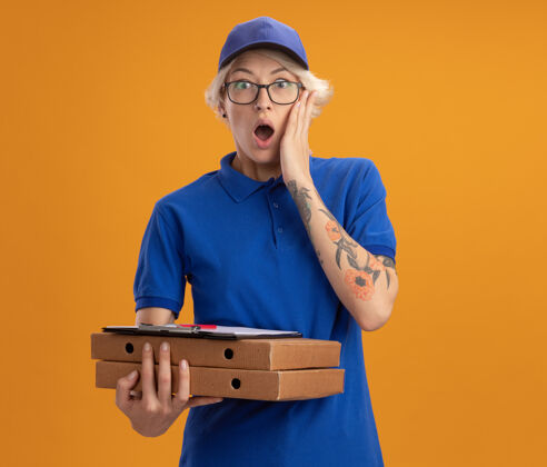 披萨身穿蓝色制服 戴着眼镜 拿着比萨饼盒和剪贴板的年轻送货员在橙色的墙上惊讶不已拿着穿年轻人