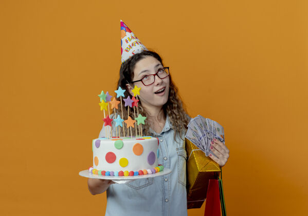 蛋糕看着戴着眼镜 戴着生日帽 拿着生日蛋糕 带着礼包的年轻女孩 盒子和钱被隔离在橙色的背景上拿着年轻人包