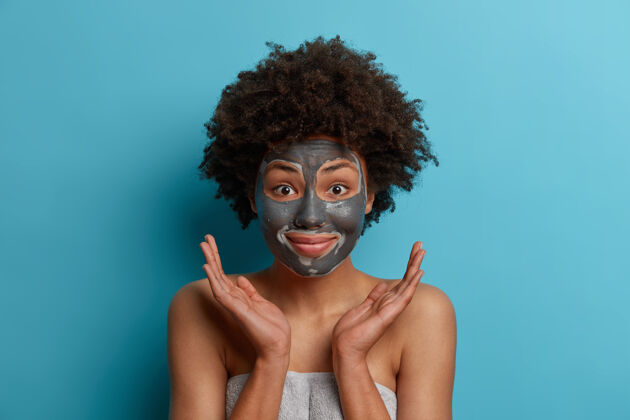 肖像积极快乐的黑皮肤非裔美国妇女使用粘土面膜 美容 护理皮肤 手掌侧放在脸上 站在毛巾包裹 模型室内卫生女士清洁美容