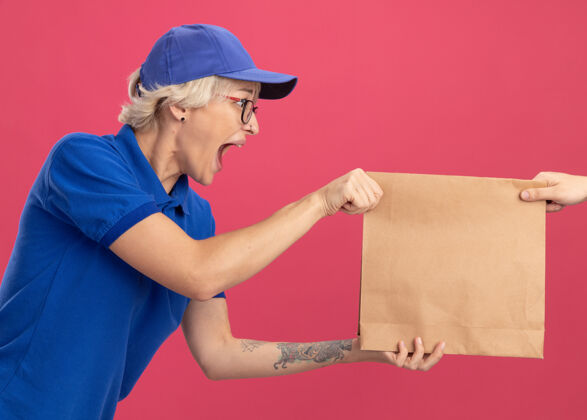 包装身穿蓝色制服 戴着帽子的年轻送货员站在粉色的墙上 一边接受纸包裹 一边兴奋不已兴奋站着帽子