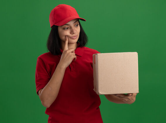 帽子年轻的送货员身穿红色制服 戴着帽子 手里拿着一个纸板箱 站在绿色的墙上困惑地看着它送货年轻拼图