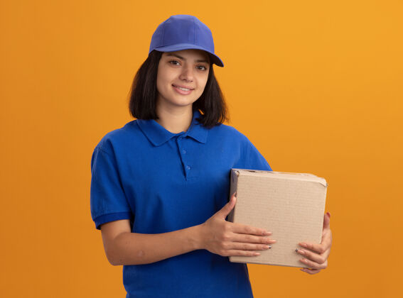 制服身着蓝色制服 头戴鸭舌帽 手拿纸板箱 友好微笑的年轻送货女孩站在橙色的墙上微笑抱着帽子