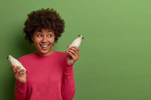 女人室内拍摄的快乐女人喝有机杏仁奶 在瓶子里摆出健康素食的姿势 拥有健康的生活方式 微笑着看着身边 穿着玫瑰色的套头衫 隔离在绿色的墙上提神有机美味