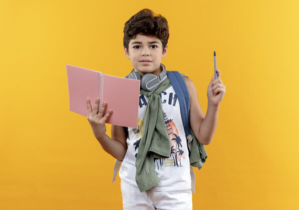 包看着摄像机 一个戴着背包和耳机的小男孩 手里拿着笔记本和笔 隔离在黄色背景上背笔抱