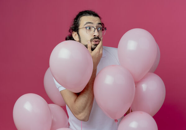 帅哥看着身边惊讶的帅哥戴着眼镜站在气球中间 把手放在脸颊上孤立的粉色背景上眼镜脸颊气球