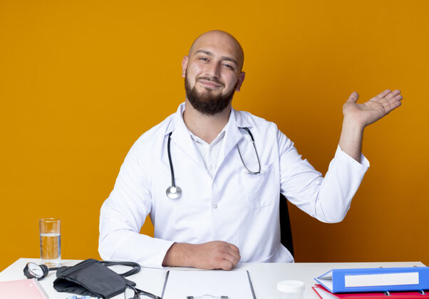 壁板高兴的年轻秃头男医生穿着医用长袍和听诊器坐在工作台上 用医疗工具点着手边隔离在橙色背景上男性年轻听诊器