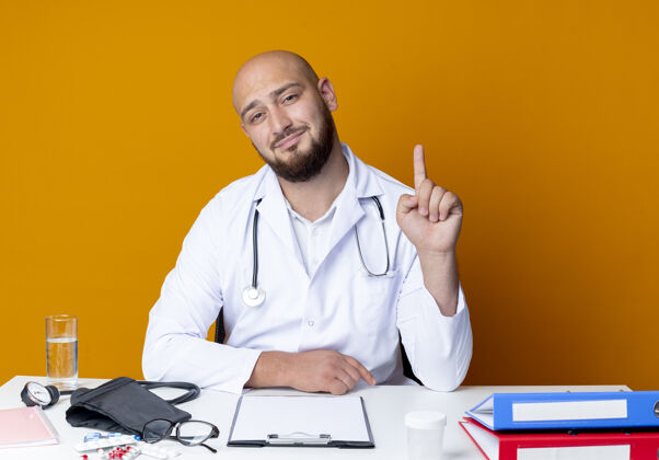 请高兴的年轻秃头男医生穿着医用长袍和听诊器坐在工作台上 用医用工具指着上面孤立的橙色背景听诊器年轻办公桌