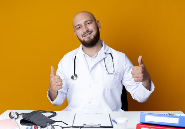 拇指年轻的秃顶男医生穿着医用长袍 手持听诊器坐在办公桌旁 手里拿着医疗工具 他的大拇指孤立地竖起在橙色的背景上工具医疗工作
