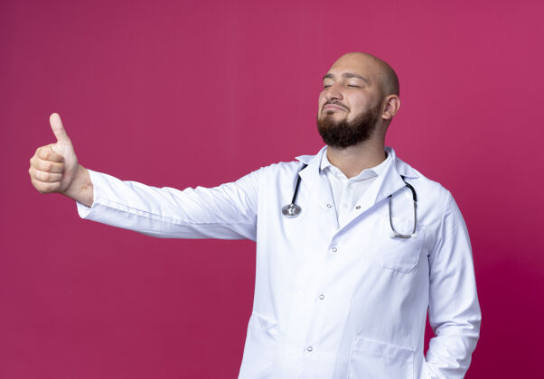 男性看着身边高兴的年轻秃头男医生穿着医用长袍和听诊器 他的大拇指孤立地竖在粉红色的背景上秃头年轻医生