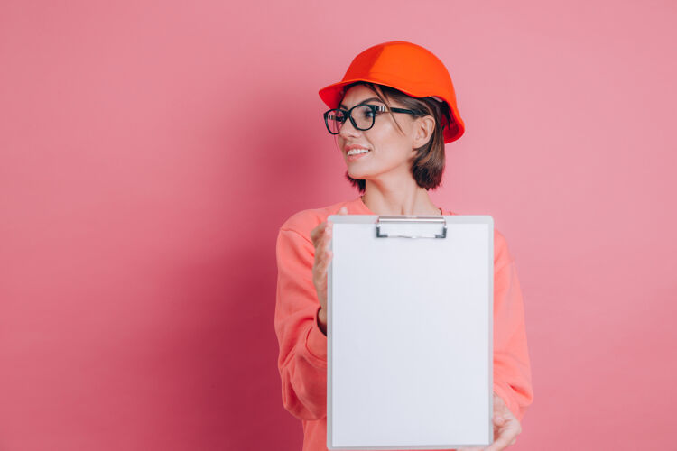 复制面带微笑的女建筑工人手持白色的空白标志牌 背景是粉色的建筑头盔女士人剪贴板