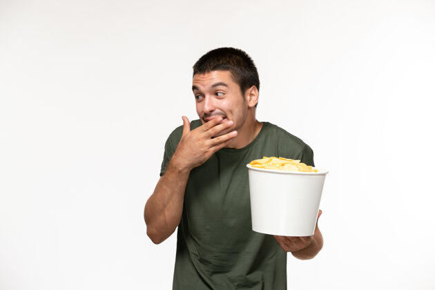 成人正面图身穿绿色t恤的年轻男子手持土豆片 在白墙上笑着孤独的电影人笑年轻的男性土豆