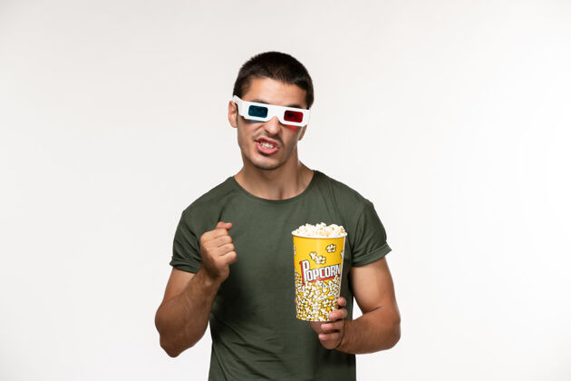 肖像正面图身着绿色t恤的年轻男性手持爆米花包戴着d墨镜在白墙上拍摄电影孤独电影院男性电影爆米花太阳镜成人
