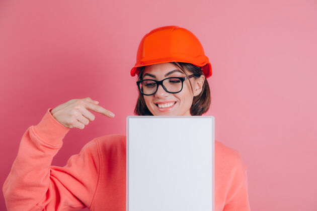 工程师面带微笑的女建筑工人拿着白色的空白标志牌 背景是粉色的戴着建筑头盔用食指指着头盔女士一个