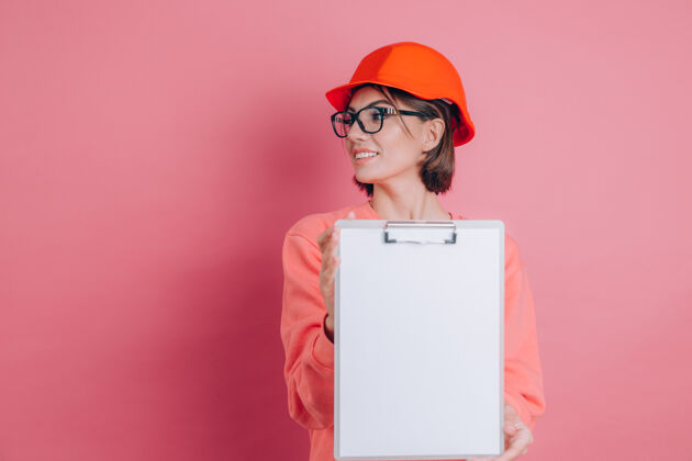 复制面带微笑的女建筑工人手持白色的空白标志牌 背景是粉色的建筑头盔女士人剪贴板