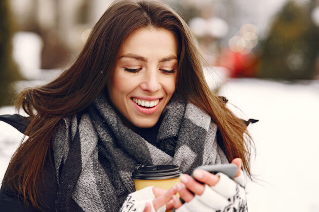 雪穿着黑色夹克喝咖啡拿着智能手机的女人的特写照片城市漂亮肖像