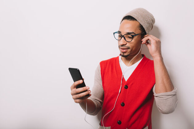 背心现代英俊的黑人男子戴着帽子和眼镜 戴着耳机听音乐 拿着灰色的智能手机休闲肖像帽子