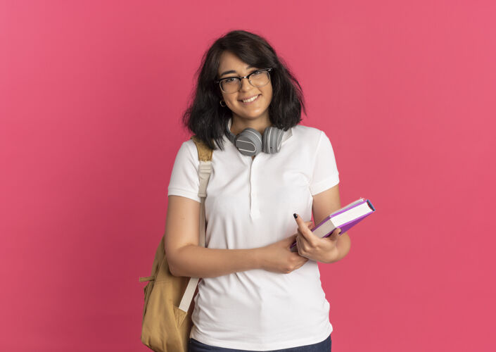 包年轻的笑容可掬的白人女生戴着眼镜 耳机 背着书包拿着粉红色的书和复印空间穿微笑粉红色