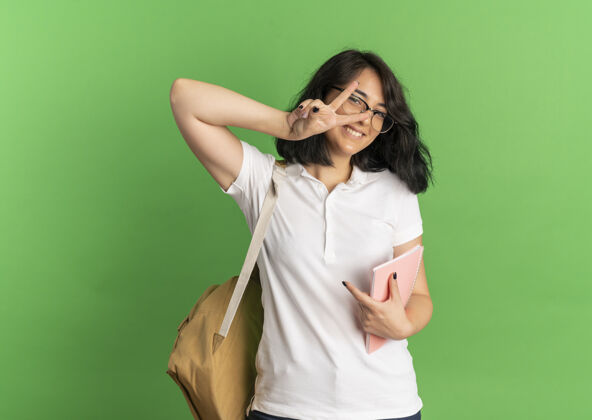 空间年轻笑容可掬的白人女学生戴着眼镜 背着书包 手势胜利 手语 手持笔记本 放在绿色的空白处手势复制包