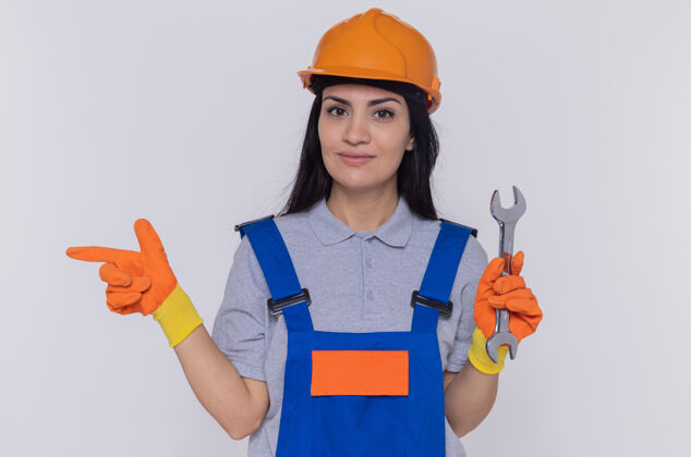 信心年轻的建筑工人妇女穿着建筑制服 戴着安全帽 戴着红色手套 拿着扳手 食指指向一边 微笑着自信地站在白色的墙上制服头盔目录