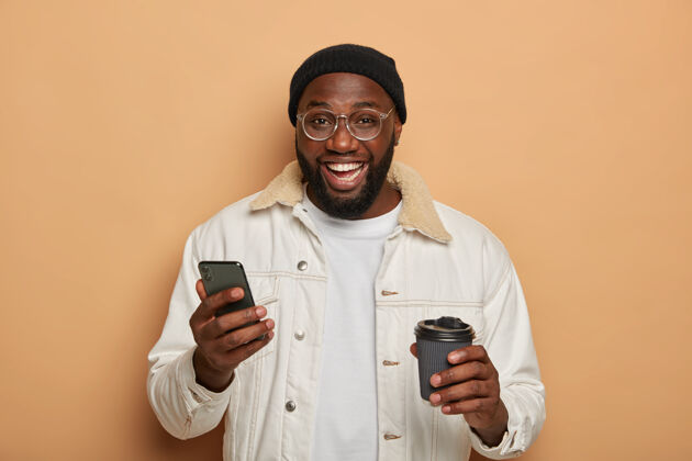 帽子黑胡子的成年男子穿着时髦的衣服 用现代智能手机上网聊天电话男人打字
