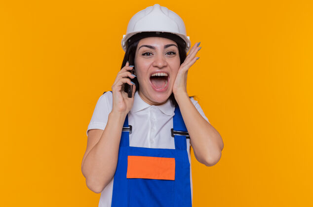疯狂身穿施工制服 戴着安全帽的年轻建筑工人站在橙色的墙上 一边用手机交谈 一边兴奋得发疯建筑女人兴奋
