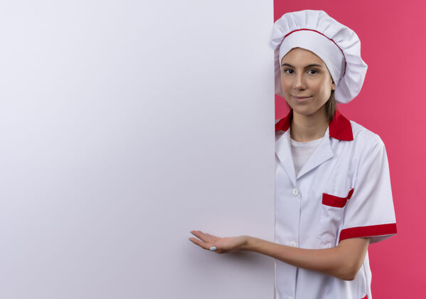 复制身着厨师制服的年轻高加索厨师女孩站在身后 指着粉色的白色墙壁 留有复印空间请年轻墙壁
