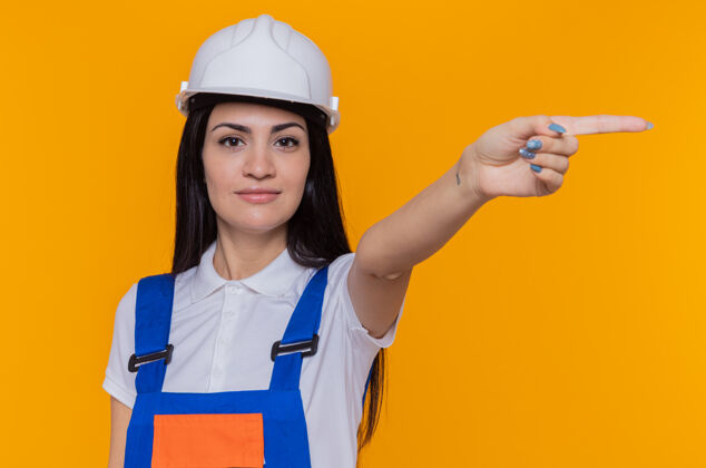 手指身穿施工制服 头戴安全帽的年轻建筑工人面带微笑 自信地用食指指着站在橙色墙上的一侧女人站建筑工
