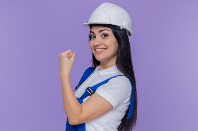 站立身穿施工制服 头戴安全帽的年轻建筑妇女微笑着自信地站在紫色的墙壁上 看着前方 露出拳头拳头年轻建筑