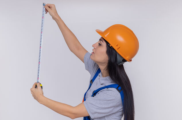 胶带身穿施工制服 头戴安全帽的年轻建筑工人站在白墙上 严肃地看着卷尺站立严肃测量