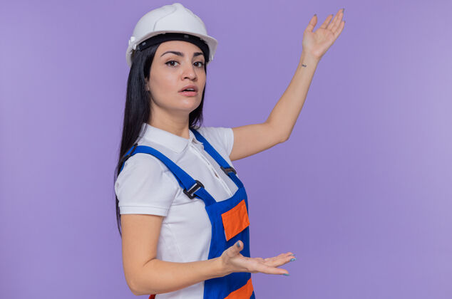 站立身穿建筑制服 头戴安全帽的年轻建筑女工人 面带严肃的表情 手臂站在紫色的墙上手臂制服什么