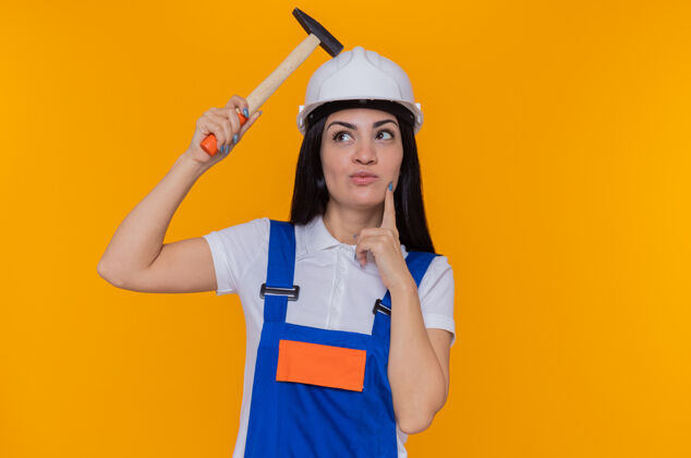 女人身穿施工制服 头戴安全帽 手持铁锤 脸上带着沉思表情的年轻建筑妇女站在橙色的墙上思考沉思站着脸