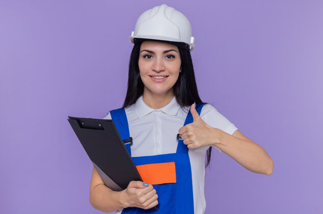 制服年轻的建筑女工穿着建筑制服 戴着安全帽拿着剪贴板显示拇指安全