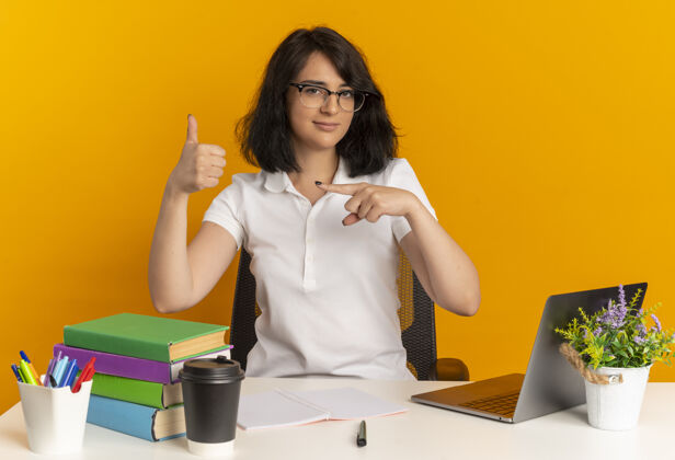 女学生年轻自信漂亮的高加索女学生戴着眼镜坐在书桌旁 手里拿着学习用具 竖起大拇指 指着旁边的橘黄色 留着复印空间年轻工具桌子