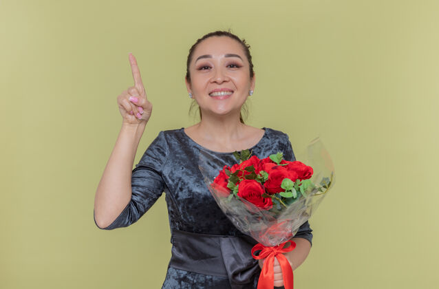 欢呼快乐的亚洲女人捧着一束红玫瑰微笑着露出食指有新想法站在绿色的墙上庆祝国际妇女节快乐微笑站立