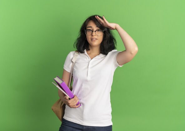 复制年轻焦急漂亮的白人女学生戴着眼镜 背着书包 手放在头上 手里拿着书 放在绿色的书架上 留着复印空间焦虑眼镜背